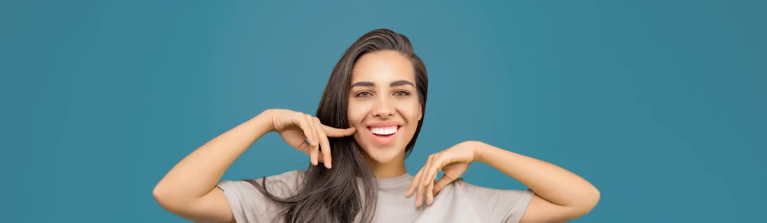 Dentist Avondale AZ Whitening A woman standing in front of a blue wall, pointing at her smile.